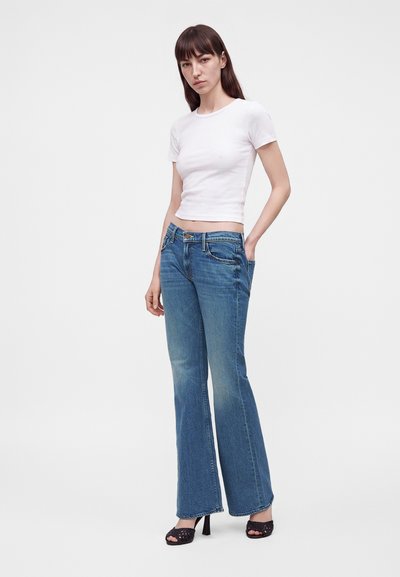 Νεαρή γυναίκα που φοράει άσπρο cropped t-shirt, μπλε καμπάνα τζιν και μαύρα σανδάλια με τακούνι, στέκεται μπροστά σε ένα απλό φωτεινό φόντο.