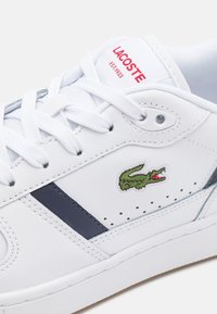 Hvit skinnsneaker med marineblå detaljer, strukturerte lisser, perforerte elementer og brodert grønn krokodillelogo på siden.