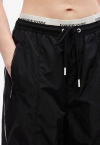 Κοντινό πλάνο ατόμου που φοράει μαύρο παντελόνι με σκοινί στη μέση, με φερμουάρ στην πλαϊνή τσέπη και ορατή λευκή ζώνη μέσης με την ετικέτα "tommy jeans."