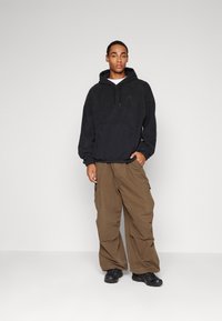 Mlad moški v črni Nike kapuci, rjavih cargo hlačah in črnih športnih copatih, stoji pred enobarvno svetlo ozadje.