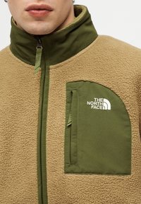 Primo piano di una persona che indossa una giacca in pile color beige con colletto verde oliva e tasca sul petto con il logo The North Face.