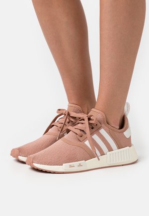 Person, die hellbraune gestrickte Sneakers mit weißen Adidas-Streifen und cremefarbenen Sohlen trägt und auf einer weißen Oberfläche steht.