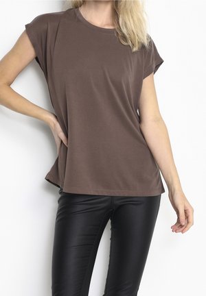 Vrouw die een loszittend bruin kortärmelig shirt en strakke zwarte leren broek draagt, poseert met de ene hand op de heup en de andere ontspannen neerhangend.