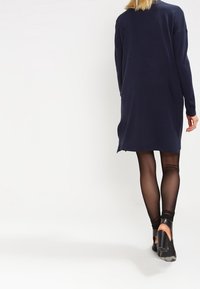 Marinblå lång klänning med lös passform, med långa ärmar och en lätt böjd nederkant, tillsammans med genomskinliga svarta strumpbyxor och högklackade skor.