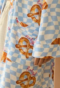 Zils un balts rūtainais audums, kas rotāts ar kliņģerīšiem un rozā krāsā rakstītu vārdu "Pretzel". Auduma struktūra izskatās viegla ar rakstainu dizainu.