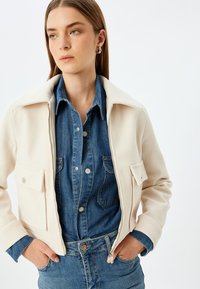 Cremfarbende Zip-Jacke mit Kragen und zwei vorderen Taschen, kombiniert mit einem blauen Jeanshemd. Weicher Stoff, lässiges Design.