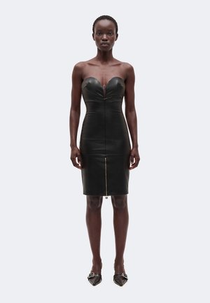 Femme portant une robe midi en cuir noir sans bretelles avec une fermeture éclair sur le devant et des talons noirs pointus avec des boucles, debout devant un fond uni.