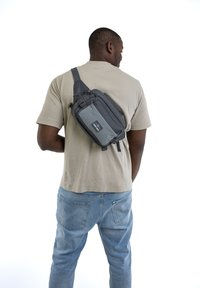 Borsa a tracolla grigia con molteplici tasche e cerniere, dotata di una tracolla resistente. Indossata sulla parte superiore del corpo, posizionata leggermente di lato.