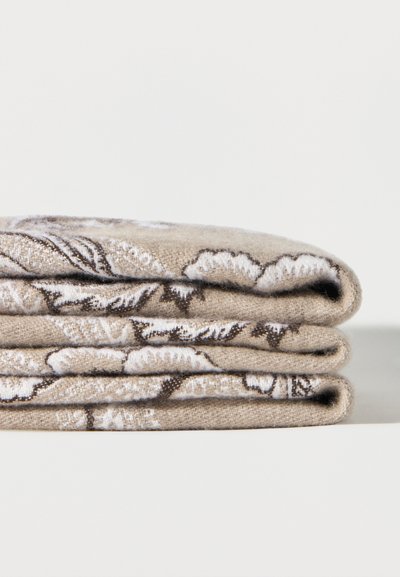 Trois pièces de tissu beige pliées avec des motifs floraux blancs et bruns empilées sur une surface blanche unie.