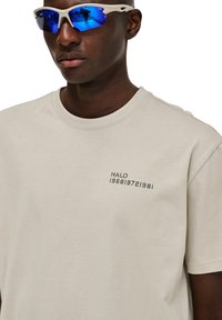 Beżowa bawełniana koszulka z krótkim rękawem, okrągłym dekoltem, z nadrukowanym napisem "HALO 196819721981" na lewej przedniej stronie.