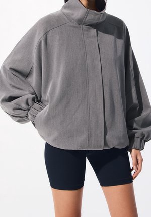 Personne portant une veste grise ample zippée avec des manches froncées et un short noir ajusté, debout devant un fond blanc uni.