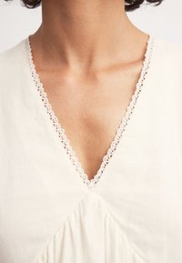 Crème-kleurig kledingstuk met een V-hals, voorzien van een decoratieve kanten afwerking en geplooide textuur onder de halslijn.