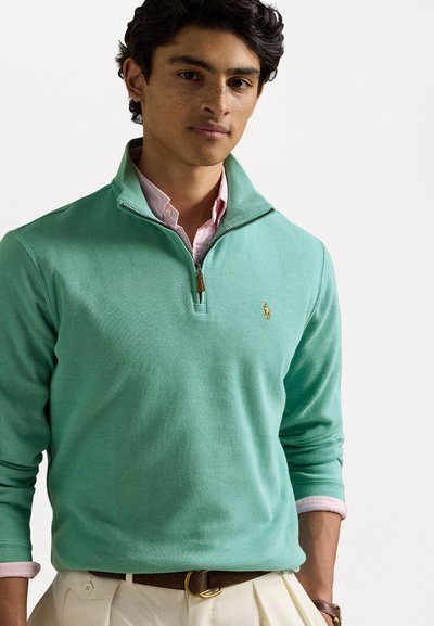 Ανοιχτό πράσινο ζακέτα με φερμουάρ φτιαγμένη από υφασμάτινη υφή, με μικρό κεντητό λογότυπο και τοποθετημένη πάνω από ένα ροζ πουκάμισο με γιακά.