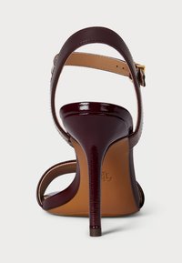 Σκούρο μπορντώ παπούτσι γόνατου με ψηλό τακούνι, λουρί στον αστράγαλο και χρυσό κούμπωμα, εμφανιζόμενο από πίσω σε λευκό φόντο.