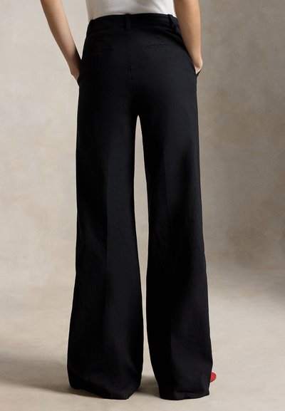 Άτομο που φοράει μαύρο παντελόνι με φαρδιά μπατζάκια και λευκό τοπ που έχει μπει μέσα στο παντελόνι, στέκεται με τα χέρια στις τσέπες μπροστά σε ουδέτερο φόντο.