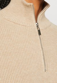 Gros plan sur un pull en maille côtelée beige avec une demi-fermeture éclair argentée et un col haut porté par une personne aux cheveux tressés foncés.