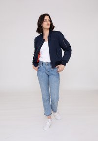 Mørkeblå bomberjakke med lynlås foran og ribbet manchetter, med et orange foer og sidelommer, parret med lyseblå jeans og hvide sneakers.