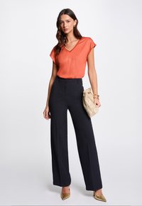 Oranje metallic V-hals blouse gecombineerd met hoge zwarte brede broek. Accessoires omvatten een gouden metallic clutch en hakken.