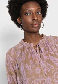 Blusa em tecido castanho claro com um padrão floral em lavanda, decote com babados e detalhe em forma de olho-chave com fecho de laço.
