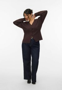 Brauner V-Ausschnitt-Cardigan mit langen Ärmeln und vorderen Knöpfen, kombiniert mit dunkelblauen, hoch taillierten Jeans. Glatte Textur, lässiger Schnitt.