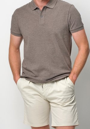 Homme portant un polo marron à manches courtes et un short beige, les mains dans les poches, devant un fond uni.