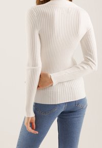 Biały prążkowany długi rękaw z dopasowanym krojem; posiada okrągły dekolt i fakturowaną tkaninę. Połączony z niebieskimi obcisłymi jeansami.