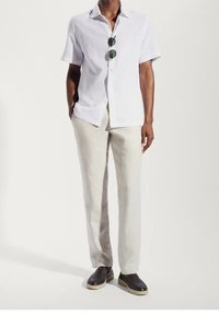 Camicetta bianca a maniche corte in lino, pantaloni in lino beige chiaro e scarpe nere senza lacci con suola chiara, accessoriate con occhiali da sole verdi.