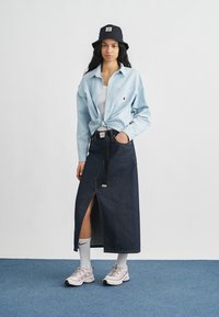 Ανοιχτό μπλε πουκάμισο με σχέδιο, δεμένο στη μέση, μακρύ φούστα από σκουρόχρωμο τζην με σκίσιμο, λευκό μπλουζάκι από μέσα και μαύρο καπέλο τύπου bucket.