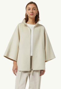 Cappotto in lana color crema con zip frontale, maniche ampie e colletto alto; presenta tasche laterali e una finitura morbida e testurizzata.