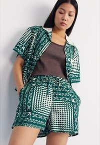 Costum format din două piese, cu un model verde, care include o cămașă cu mânecă scurtă, cu accente albe, și pantaloni scurți cu talie elastică, ambele având un design geometric.