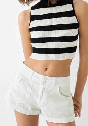 Kvinde iført sort og hvid stribet ærmeløs crop top med hvide højtaljede shorts med frontlommer og dobbelte knapper.