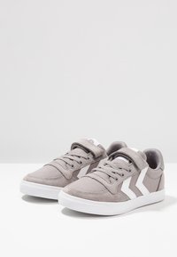 Zapatillas casuales grises con franjas de chevrón blancas, cordones y correas de velcro, colocadas una al lado de la otra sobre una superficie blanca.