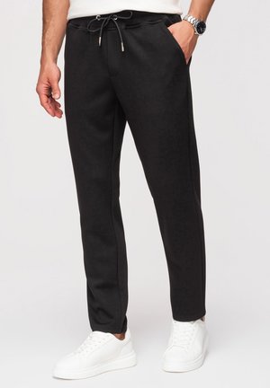 Homme portant un pantalon de jogging noir à cordon avec poches latérales et des baskets blanches, debout avec une main dans la poche.