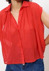 Mujer vestida con una blusa roja suelta sin mangas con botones y jeans de mezclilla azules, de pie con una mano en la cadera.