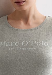 Graues, kurzärmliges T-Shirt mit einem Rundhalsausschnitt. Auf der Vorderseite befindet sich der Text „Marc O'Polo EST. IN STOCKHOLM“ in Hellgrau.