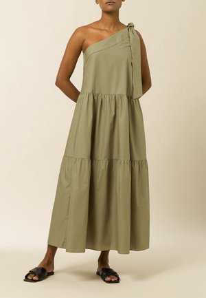 Woman wearing olive green one-shoulder tiered maxi dress with bow detail, paired with black flat slide sandals on beige background.