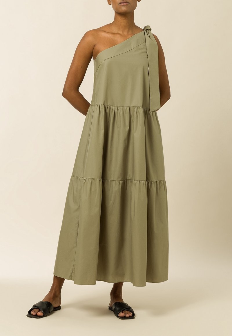 Femme portant une robe maxi olive à bretelle unique et à volants avec un détail de nœud, associée à des sandales plates noires sur fond beige.