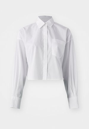 Chemise blanche à boutons, raccourcie avec des manches longues, une poche sur le côté gauche et un col classique pointu, présentée sur un fond blanc.
