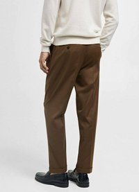 Bruine op maat gemaakte broeken van gladde stof; voorzien van een enkele achterzak met een knoop; gestyled met opgerolde manchetten en zwarte schoenen.