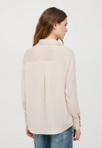 Camicia lunga beige pallido con una texture morbida e liscia, dettaglio di piega centrale sul retro e colletto classico, indossata sopra jeans in denim blu.