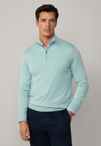 Helleblaue, Pullunder mit Reißverschluss aus weichem Strickmaterial, mit gerippten Bündchen und Saum. Darüber getragen hat es ein blau gestreiftes Hemd mit Kragen.