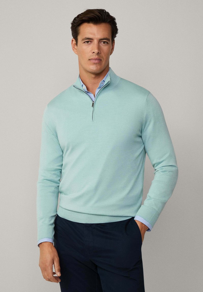 Helleblaue, Pullunder mit Reißverschluss aus weichem Strickmaterial, mit gerippten Bündchen und Saum. Darüber getragen hat es ein blau gestreiftes Hemd mit Kragen.