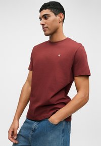 Bordový crewneck tričko s krátkymi rukávmi, vyrobené z bavlny, s malým logom na ľavej hrudi a voľným strihom. Nosené s svetlo modrými džínsami.