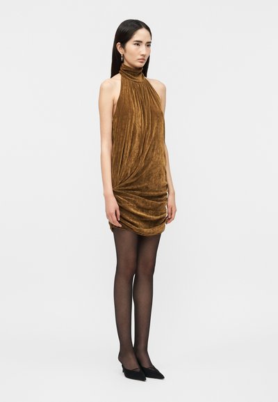 Γυναίκα που φοράει αμάνικο καφέ βελούδινο φόρεμα halter με ραφές, μαύρο διάφανο καλσόν και μαύρες μυτερές γόβες, στέκεται μπροστά σε λευκό φόν το.