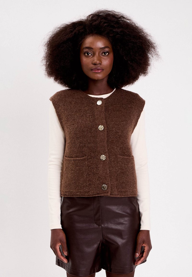 Femme avec des cheveux bouclés volumineux portant un gilet en tricot marron à boutons sur un top à manches longues blanc et un short en cuir marron foncé.