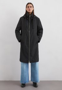 Manteau noir zippé avec capuche, doté de deux poches avant et de liens, associé à un jean bleu délavé à jambes larges et des bottes noires. Texture lisse.