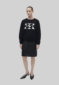 Maglione nero con una grafica bianca; indossato con una gonna di pelle nera. Il modello indossa tacchi a spillo neri e uno sfondo neutro.