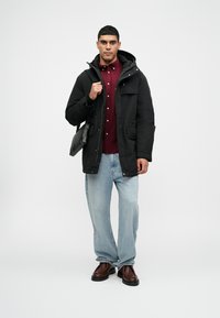 Casaco parka preto com capuz, combinado com uma camisa bordô de botões. Usado com jeans azul claro e sapatos castanhos escuros. Segura uma bolsa preta.
