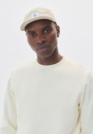 Light beige cap with a curved brim and stitched emblem featuring "M 73." Accompanied by a pale yellow, crew-neck sweatshirt.