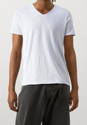 Hombre vistiendo una camiseta de manga corta con cuello en V blanca y lisa, con pantalones gris oscuro, de pie contra un fondo claro y liso.